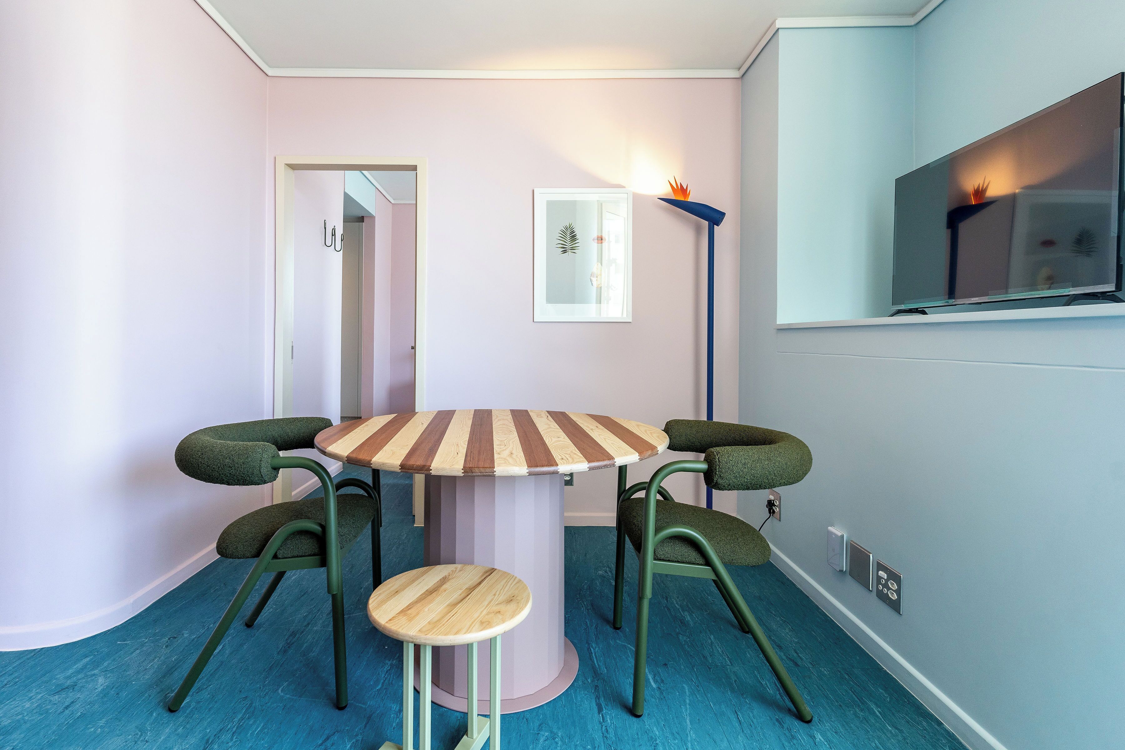 A room with a round table, four chairs, a small stool, a floor lamp, and a wall-mounted TV.