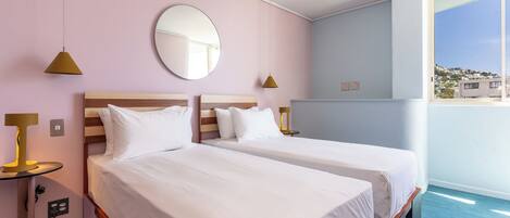 A hotel room with two beds, a round mirror, bedside lamps, a small table, and a window with a view of buildings.