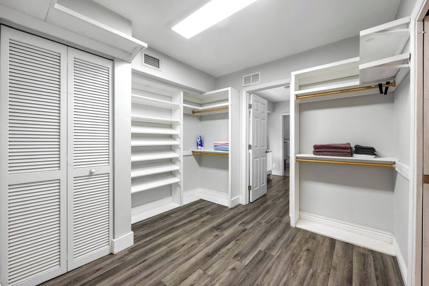 A walk-in closet with white shelving, a wooden floor, and a door leading to another room.