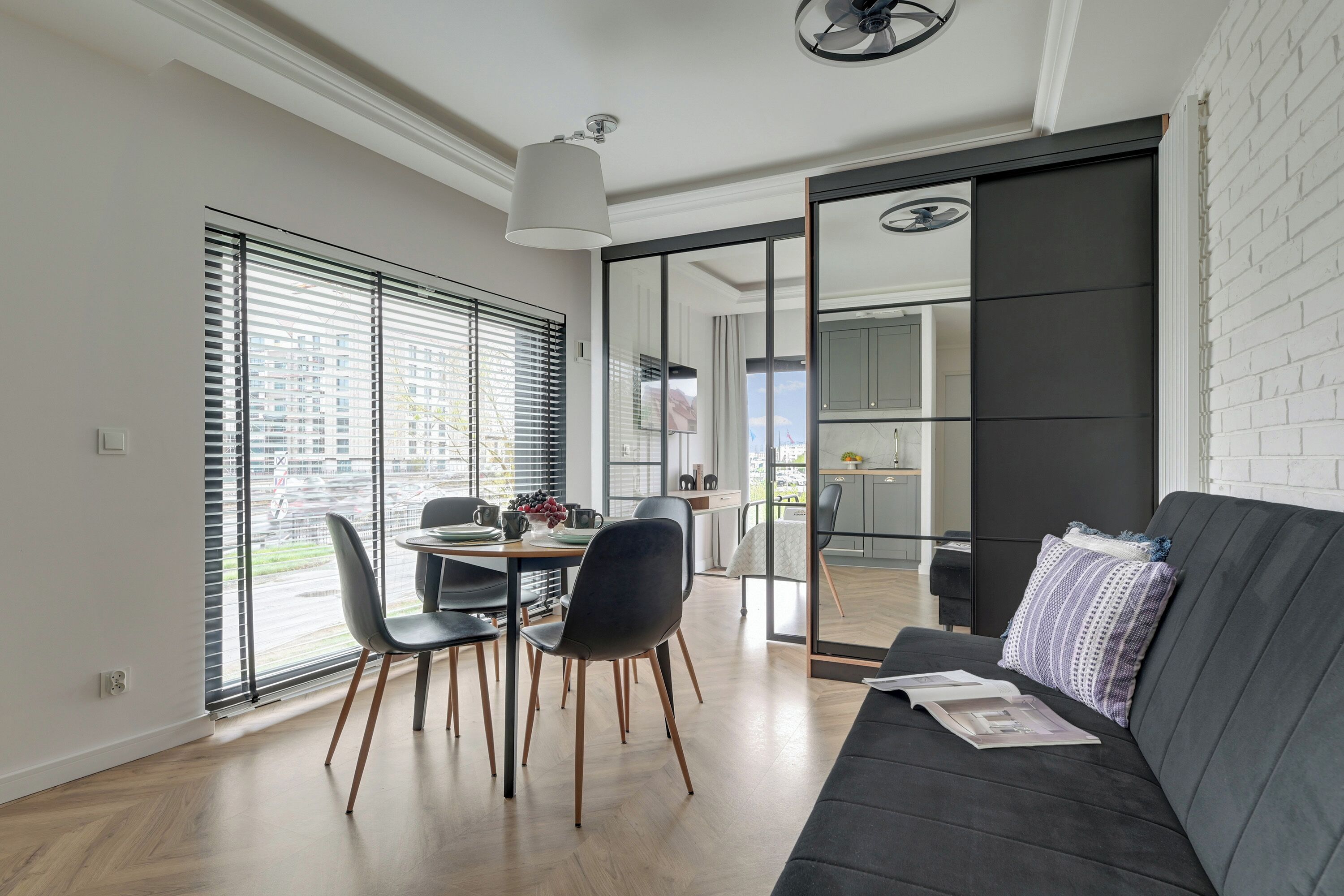 A modern living room with a dark sofa, a dining table with chairs, and a large window with blinds.