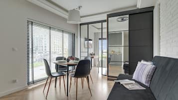 A modern living room with a dark sofa, a dining table with chairs, and a large window with blinds.