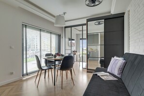 A modern living room with a dark sofa, a dining table with chairs, and a large window with blinds. - Rezydencja Wintera by Downtown Apartments (Gdansk)