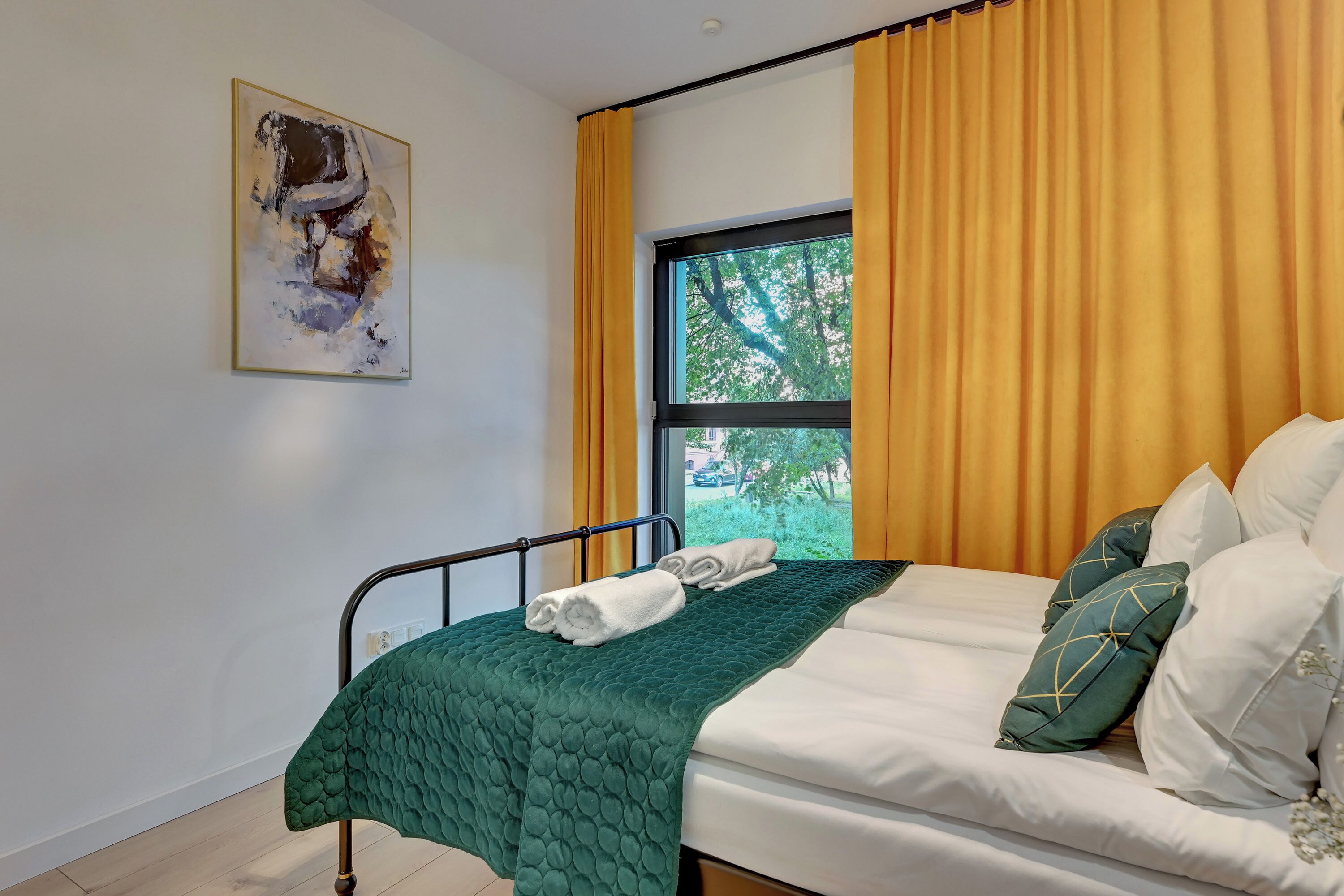 Una cama bien hecha con colcha verde, almohadas blancas y una ventana con cortinas amarillas.