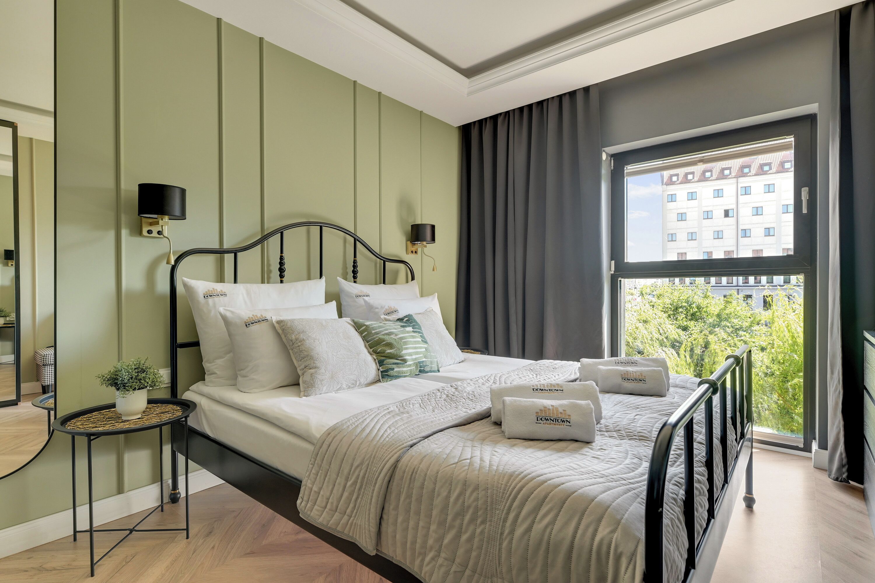 Ein ordentlich eingerichtetes Schlafzimmer mit Bett, Nachttischen, einem Fenster mit Vorhängen und Blick auf Gebäude.