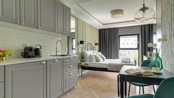 A hotel room with a bed, a desk with a chair, a dining table, a kitchen area with cabinets, and a window with a view of buildings.
