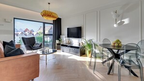 A modern living room with a sofa, a dining table, and chairs. There is a TV mounted on the wall and a large window providing a view of buildings outside.