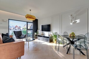A modern living room with a sofa, a dining table, and chairs. There is a TV mounted on the wall and a large window providing a view of buildings outside.