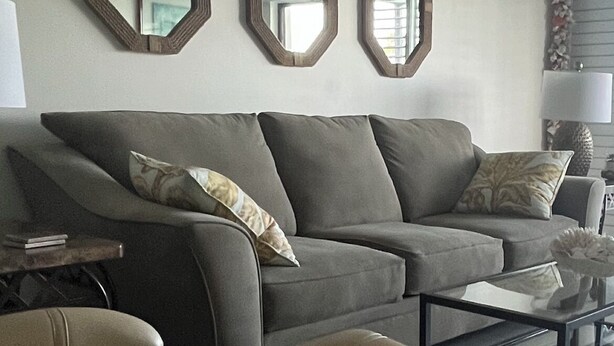 A living room with a grey sofa, two ottomans, a glass coffee table, and three wooden mirrors on the wall.