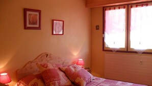 Ein Schlafzimmer mit einem Bett, zwei Nachttischlampen, einem Fenster mit Vorhängen und zwei gerahmten Bildern an der Wand.
