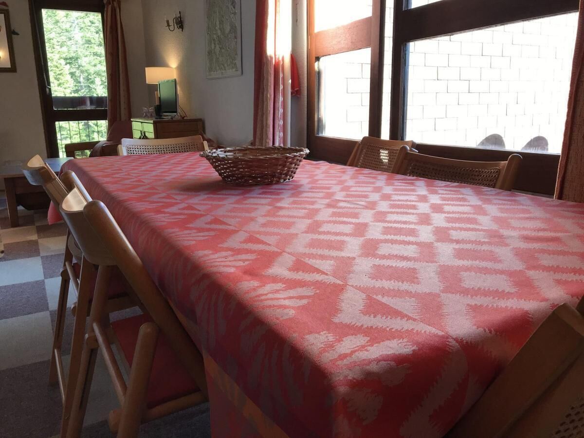A dining area with a checkered tablecloth, wooden chairs, a basket, and a window with a brick wall view.