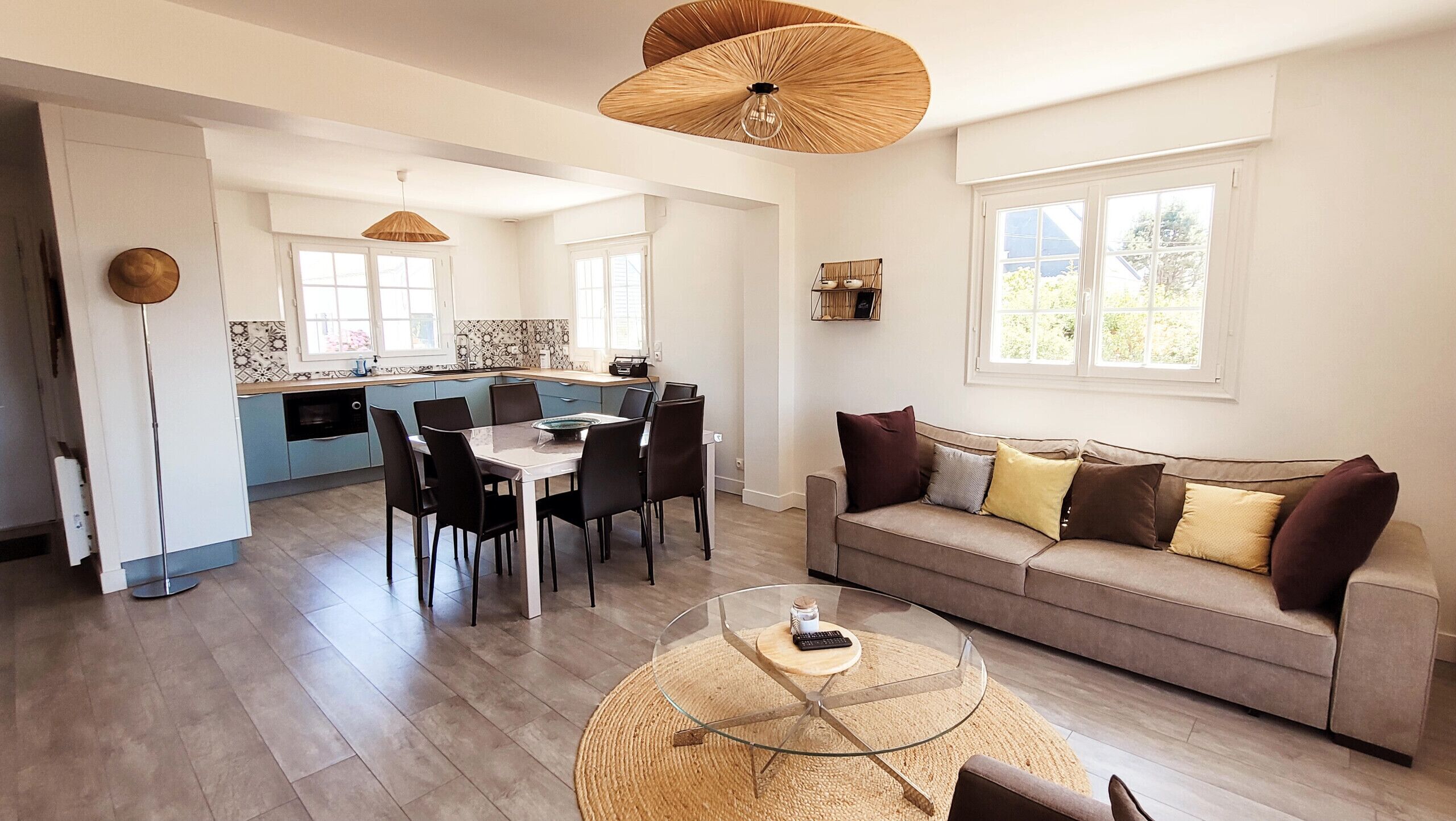 Un salon moderne avec une table basse en verre, un canapé avec des coussins, un coin repas avec des chaises et une cuisine en arrière-plan.