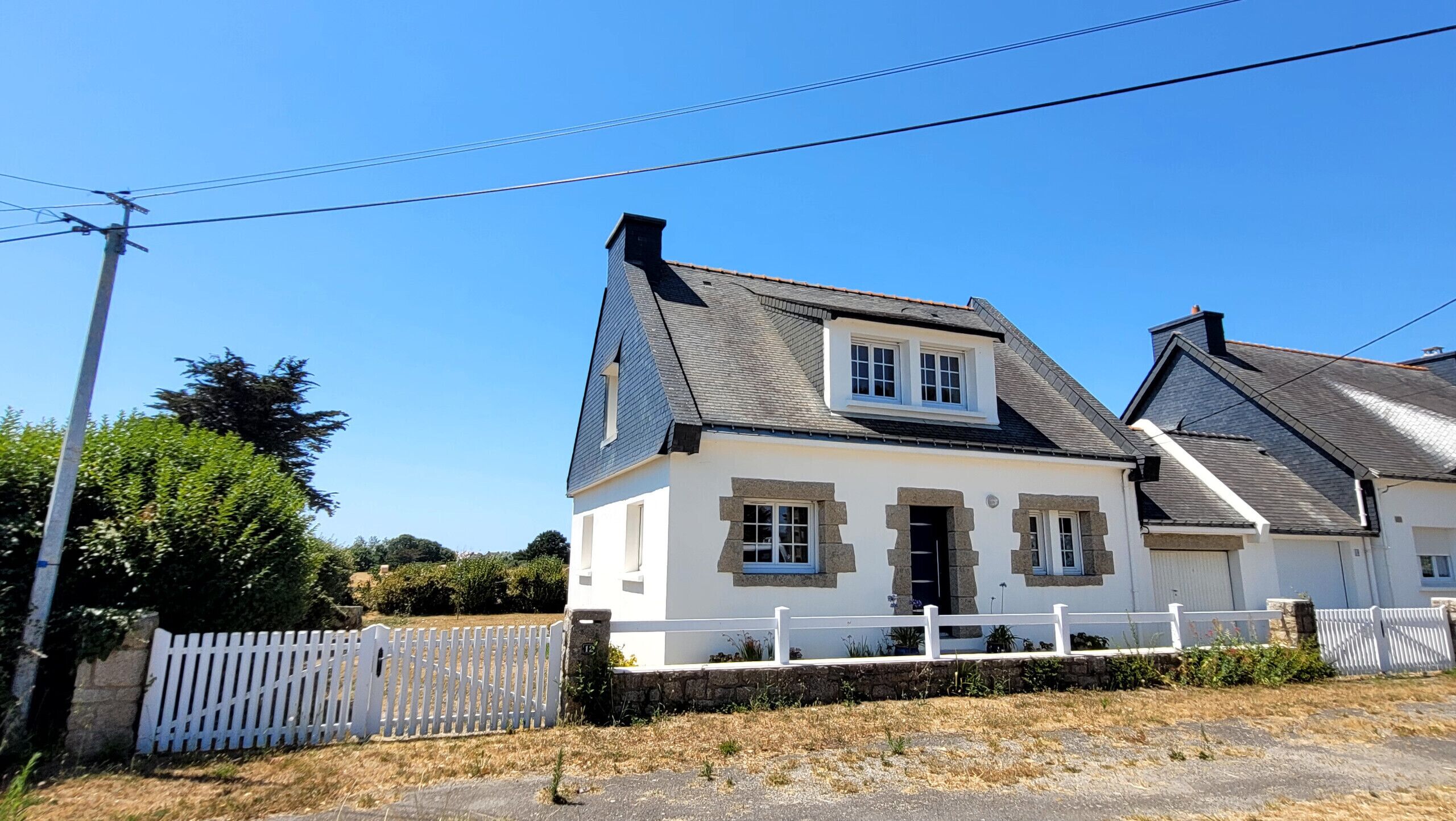 Une maison à deux étages, avec une façade blanche, des fondations en pierre et un toit à deux versants.