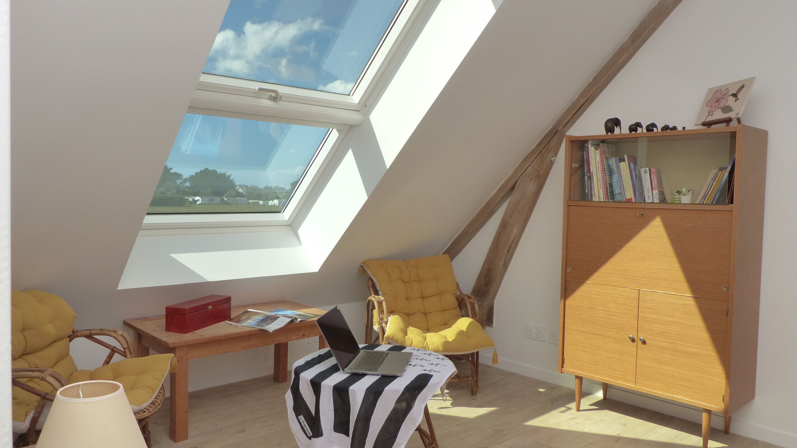 Une pièce avec un plafond en pente, des fenêtres de toit, une bibliothèque en bois, un fauteuil jaune, un bureau en bois et un ordinateur portable.