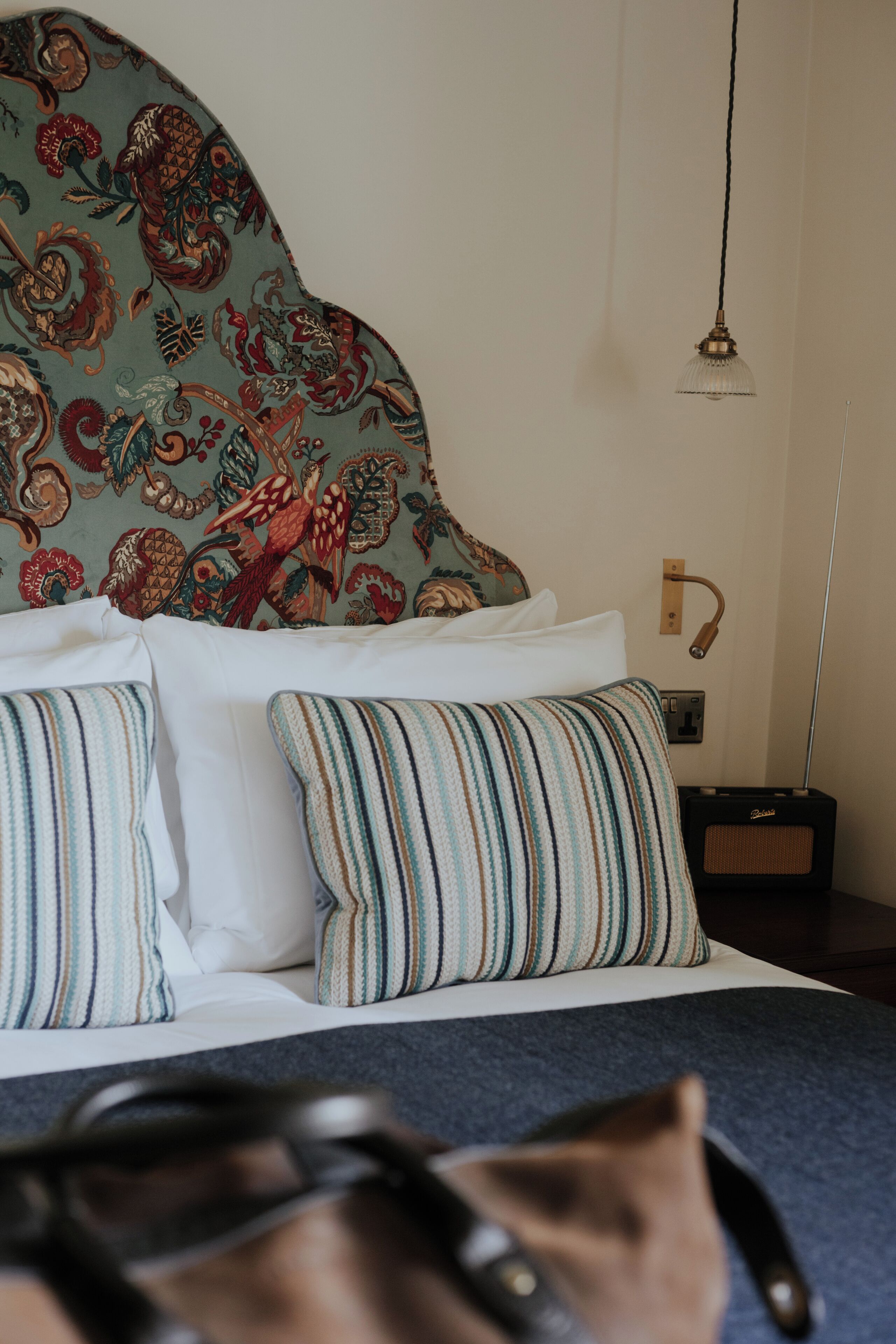 A bed with striped pillows, a patterned headboard, a hanging light, and a radio on a bedside table.