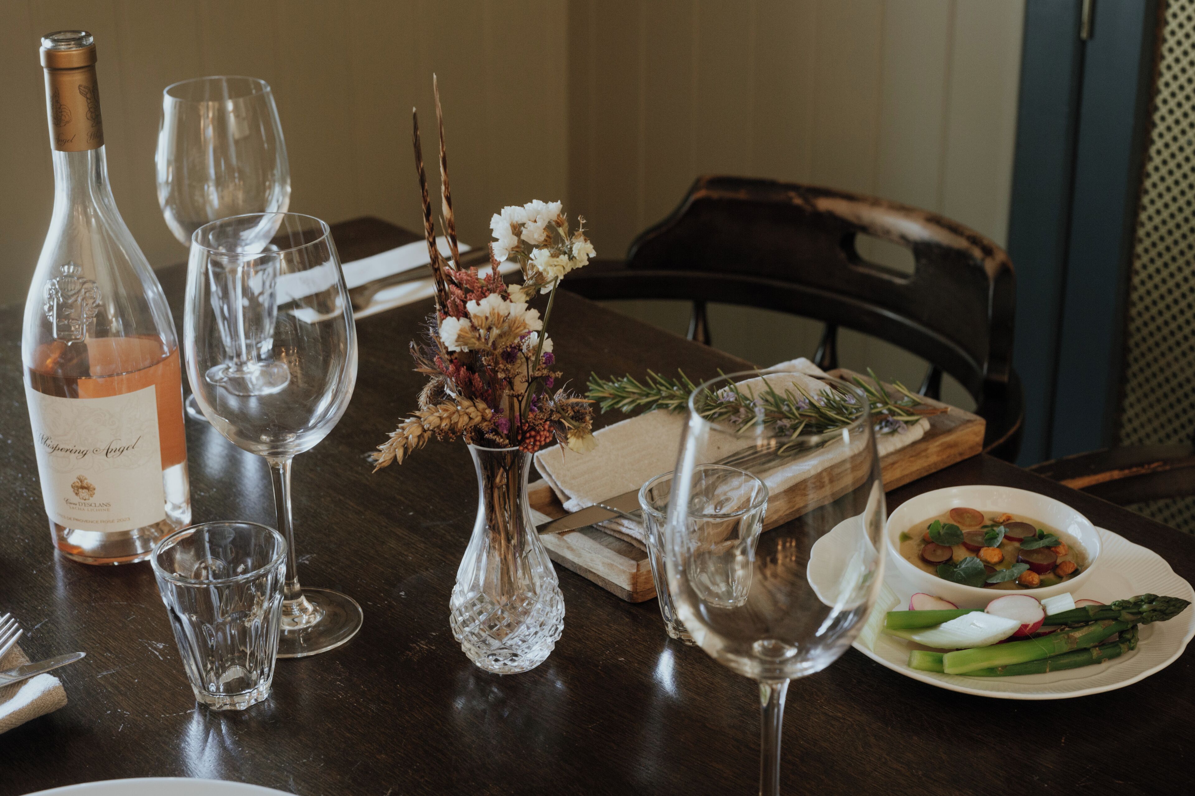 Et spisebord med en flaske vin, tomme vinglas, en vase med tørrede blomster, en træplanke med mad og en tallerken med grøntsager.