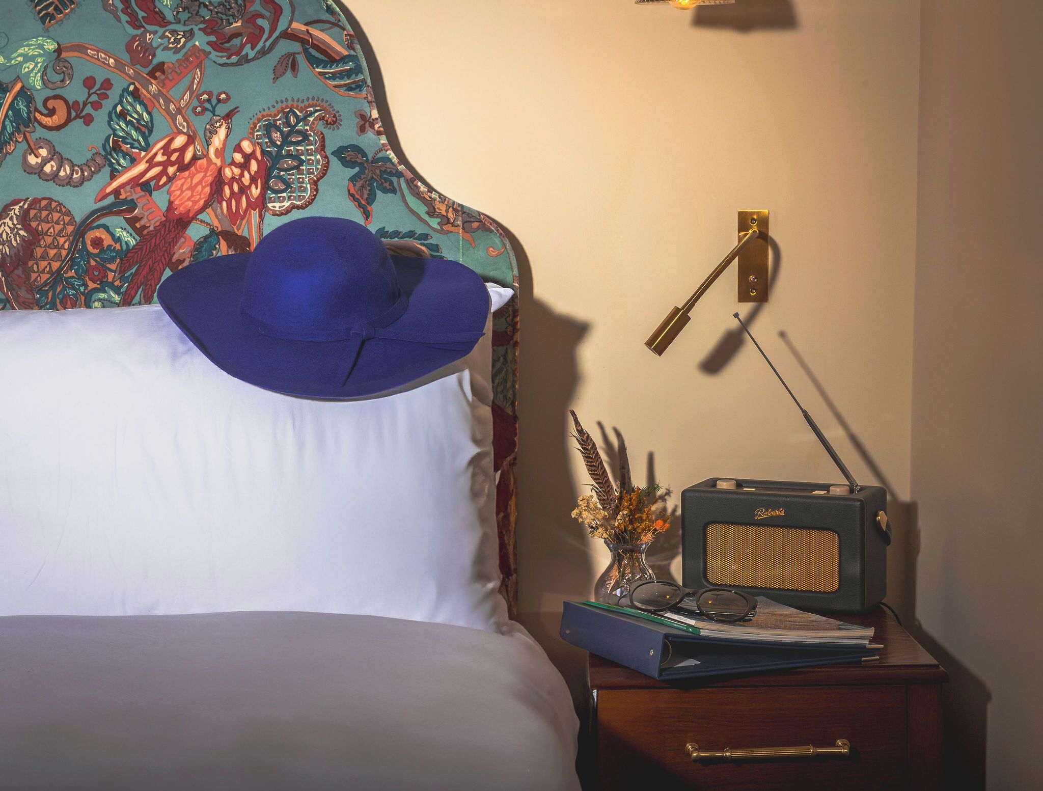 Uma cama com um chapéu azul sobre o travesseiro, um rádio em uma mesinha de cabeceira e um painel decorativo na parede.