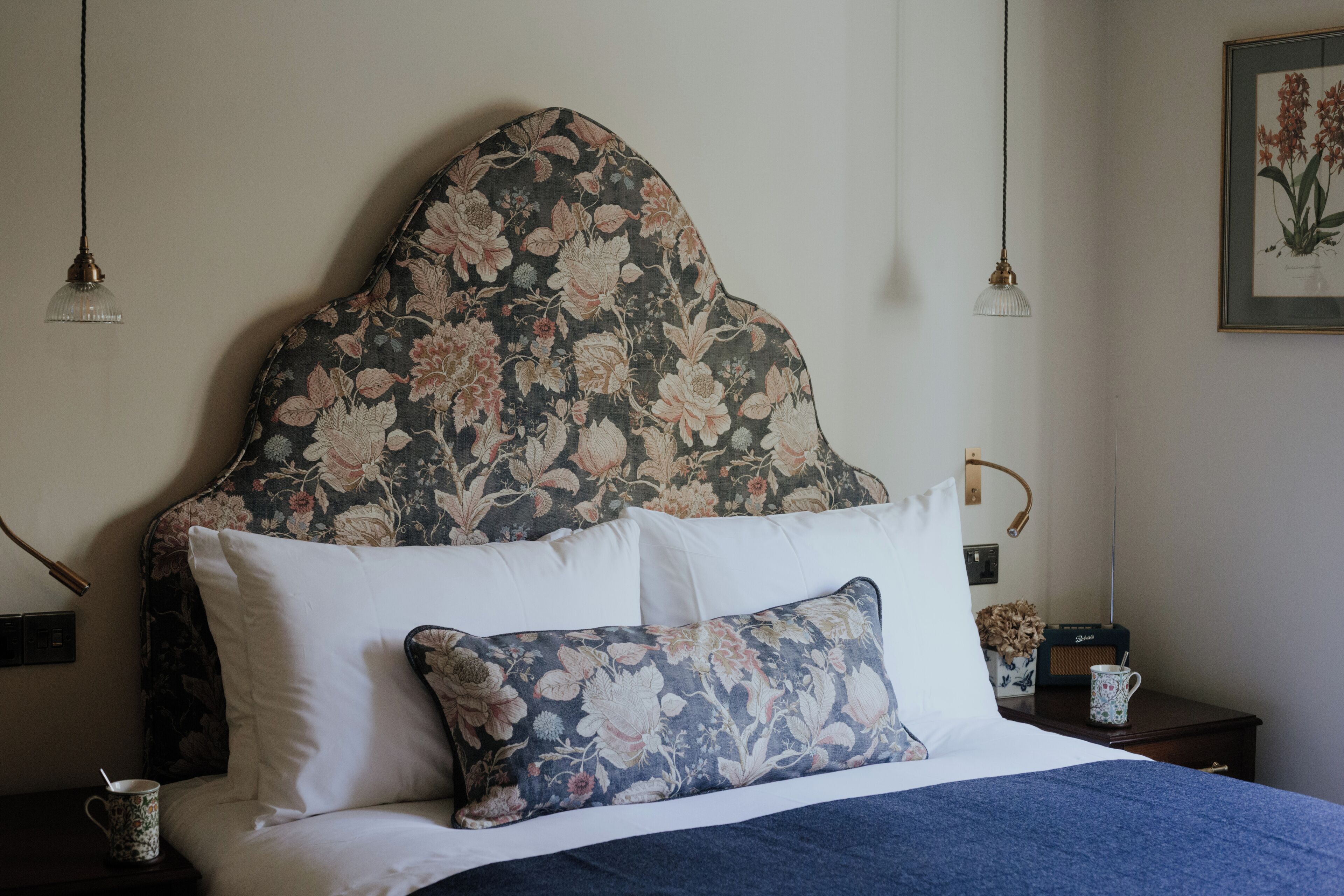 A neatly made bed with floral-patterned headboard and pillows, a bedside table with a radio and a mug, and a framed botanical print on the wall.