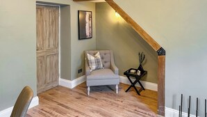 Une pièce avec une porte en bois, un fauteuil gris, une petite table sur laquelle se trouve un vase, et un plafond avec une poutre en bois.