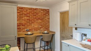 Une cuisine compacte avec un mur en briques, deux tabourets de bar, un comptoir blanc et une machine à café.