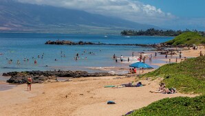 A beach with people swimming and sunbathing, a rocky outcrop, and a distant mountain range.