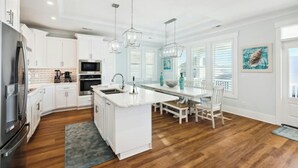 Une cuisine moderne avec des meubles blancs, un îlot central, des appareils en acier inoxydable et un coin repas comprenant une table et des chaises blanches.