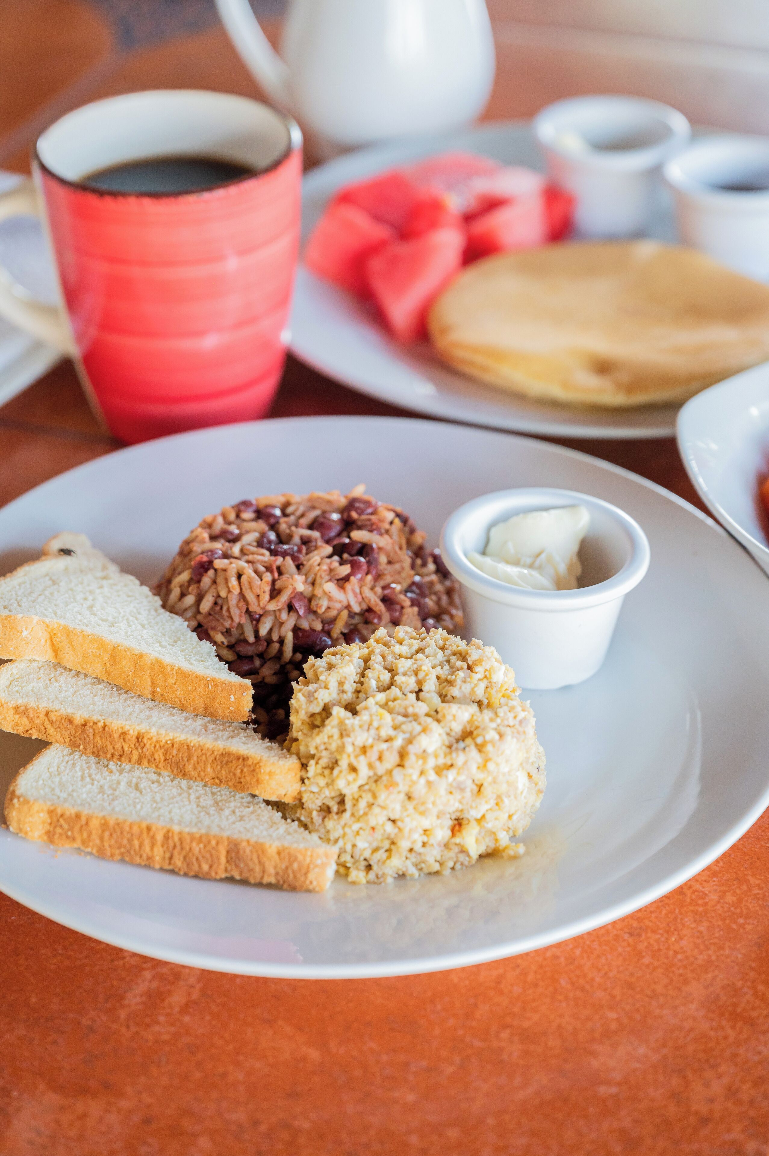 A plate with rice, beans, and toast, a red mug, and a white cup with butter.