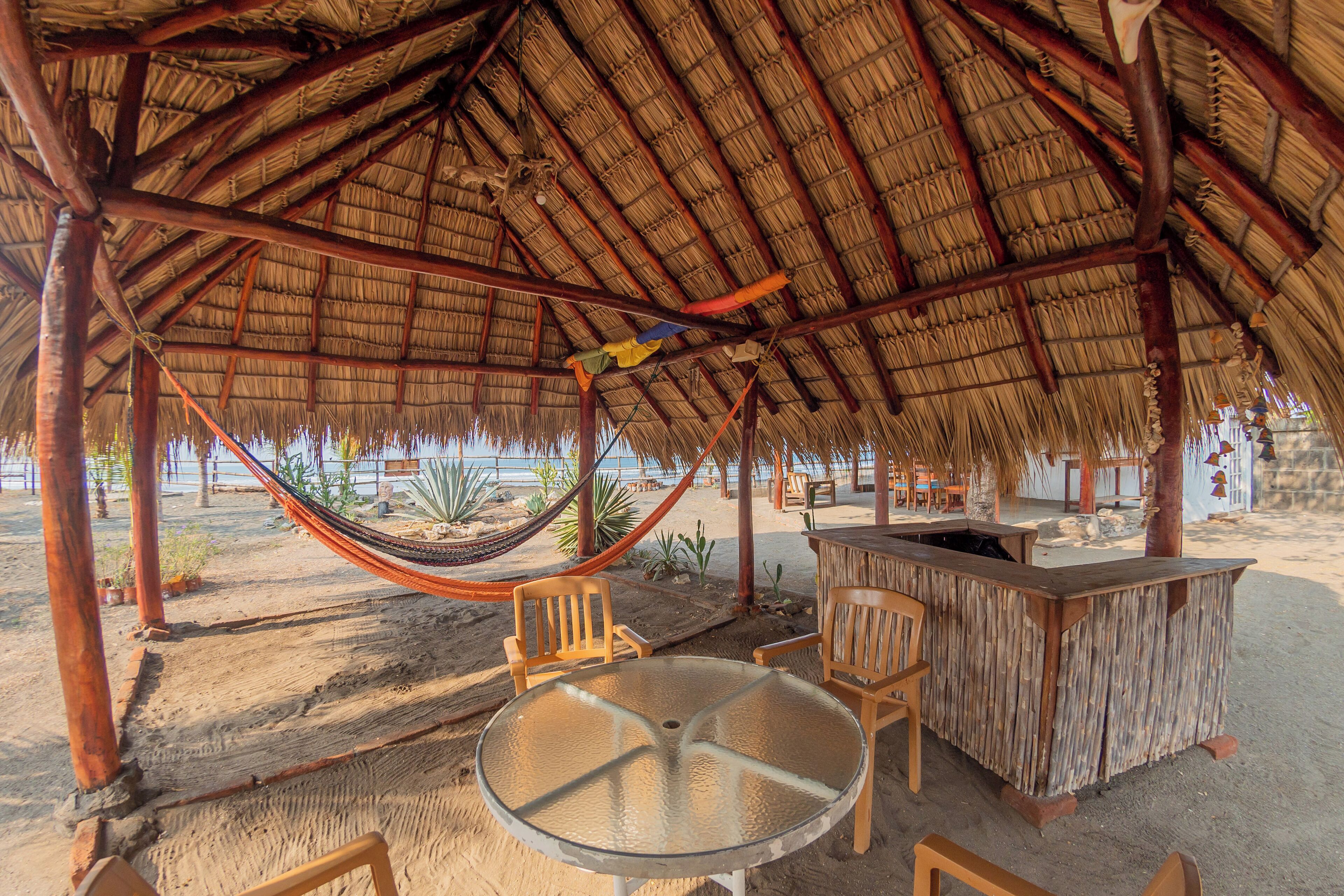 A beachside hut with a thatched roof, a glass table, wooden chairs, and a wooden bar.