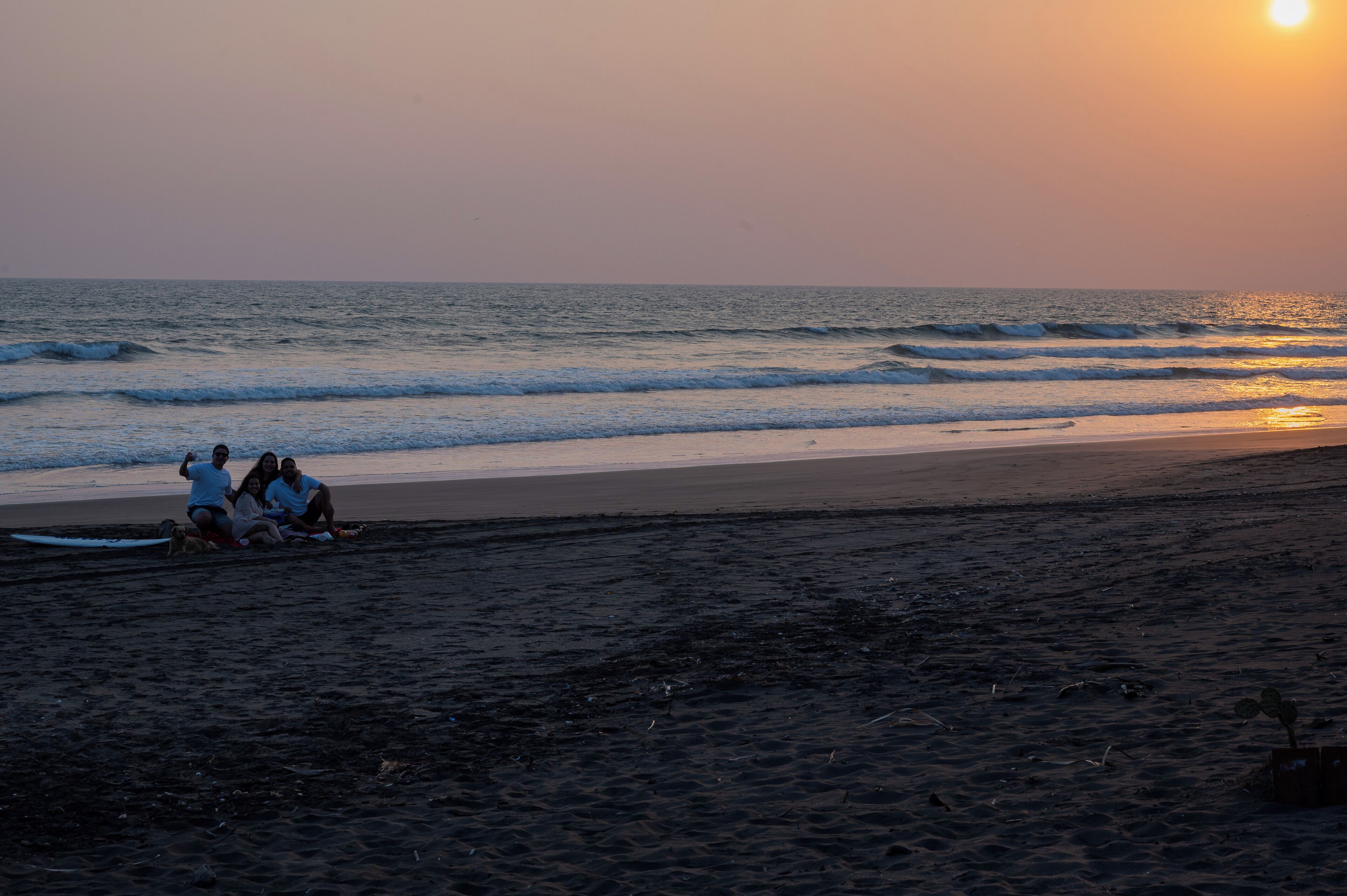 Ein Strandbild mit Sonnenuntergang, Wellen und einer Gruppe Menschen, die im Sand sitzen.