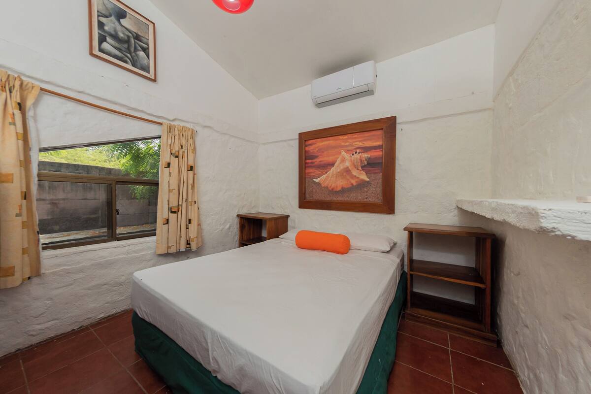 Une chambre à coucher avec un lit, une fenêtre avec des rideaux, un tableau au mur et une petite table de chevet en bois.