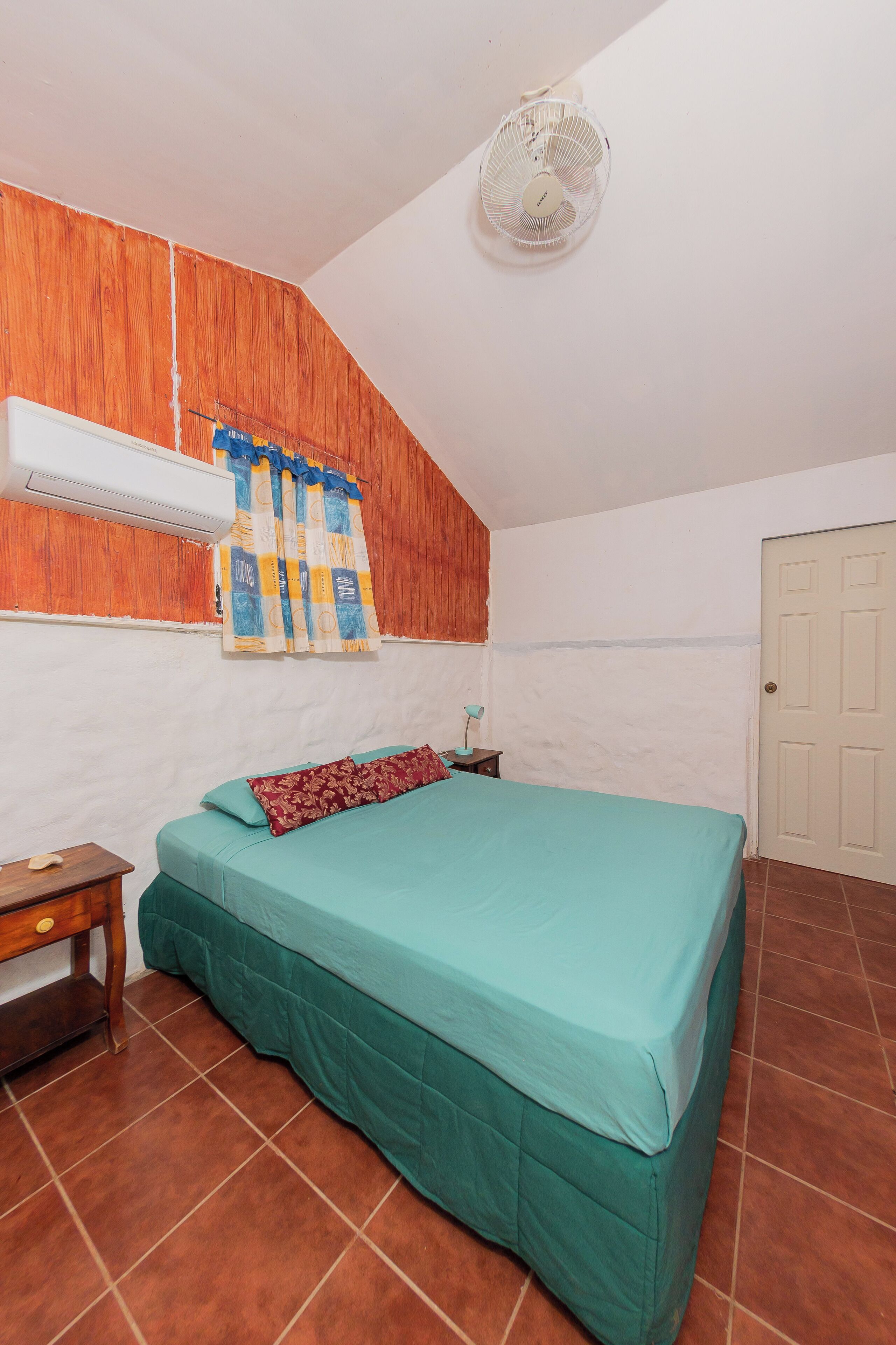 A bedroom with a bed, a nightstand, a wall-mounted air conditioner, a ceiling fan, and a door.