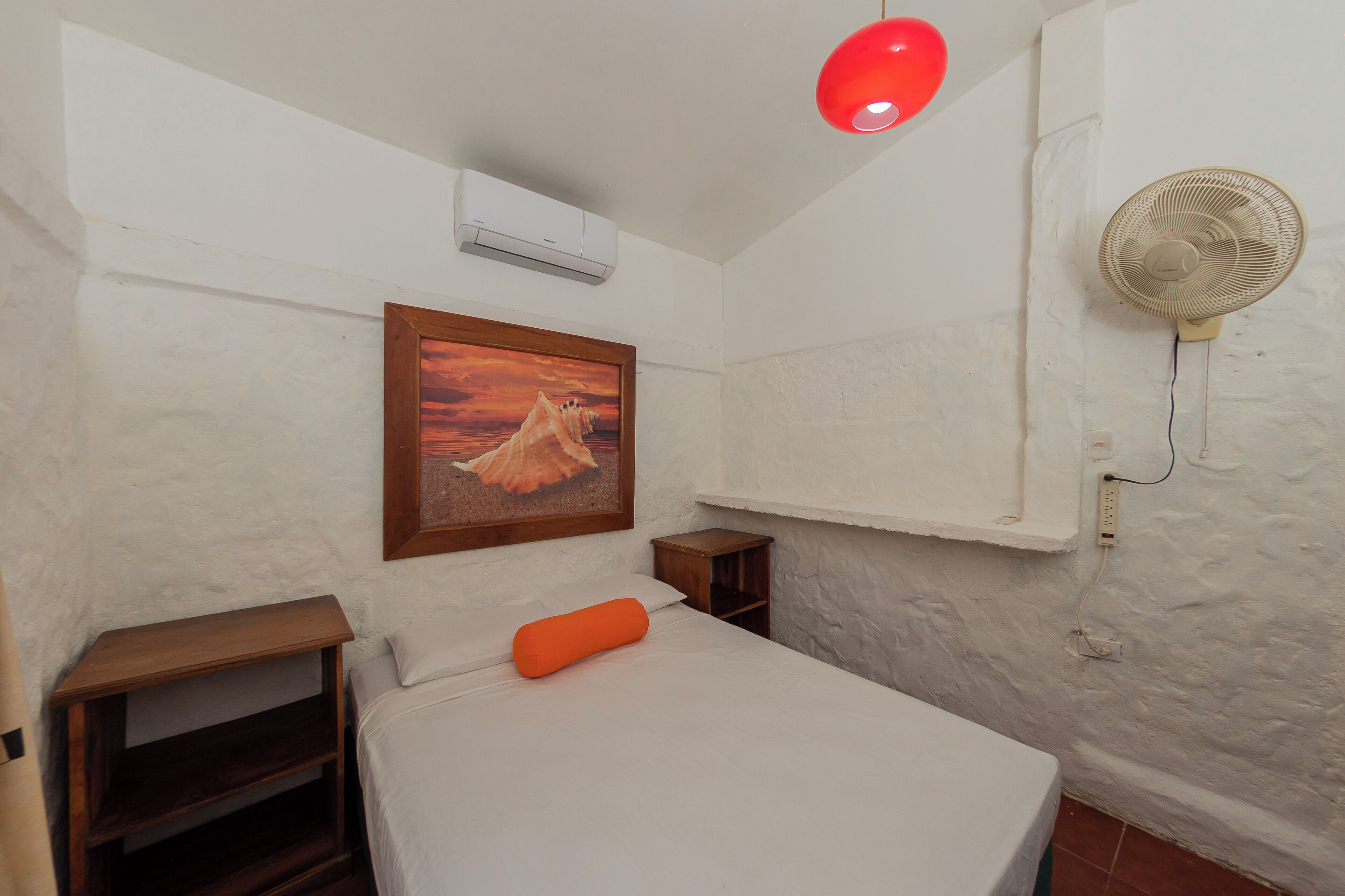 A bedroom with a bed, a small wooden nightstand, a wall-mounted fan, a red light fixture, and a framed picture on the wall.