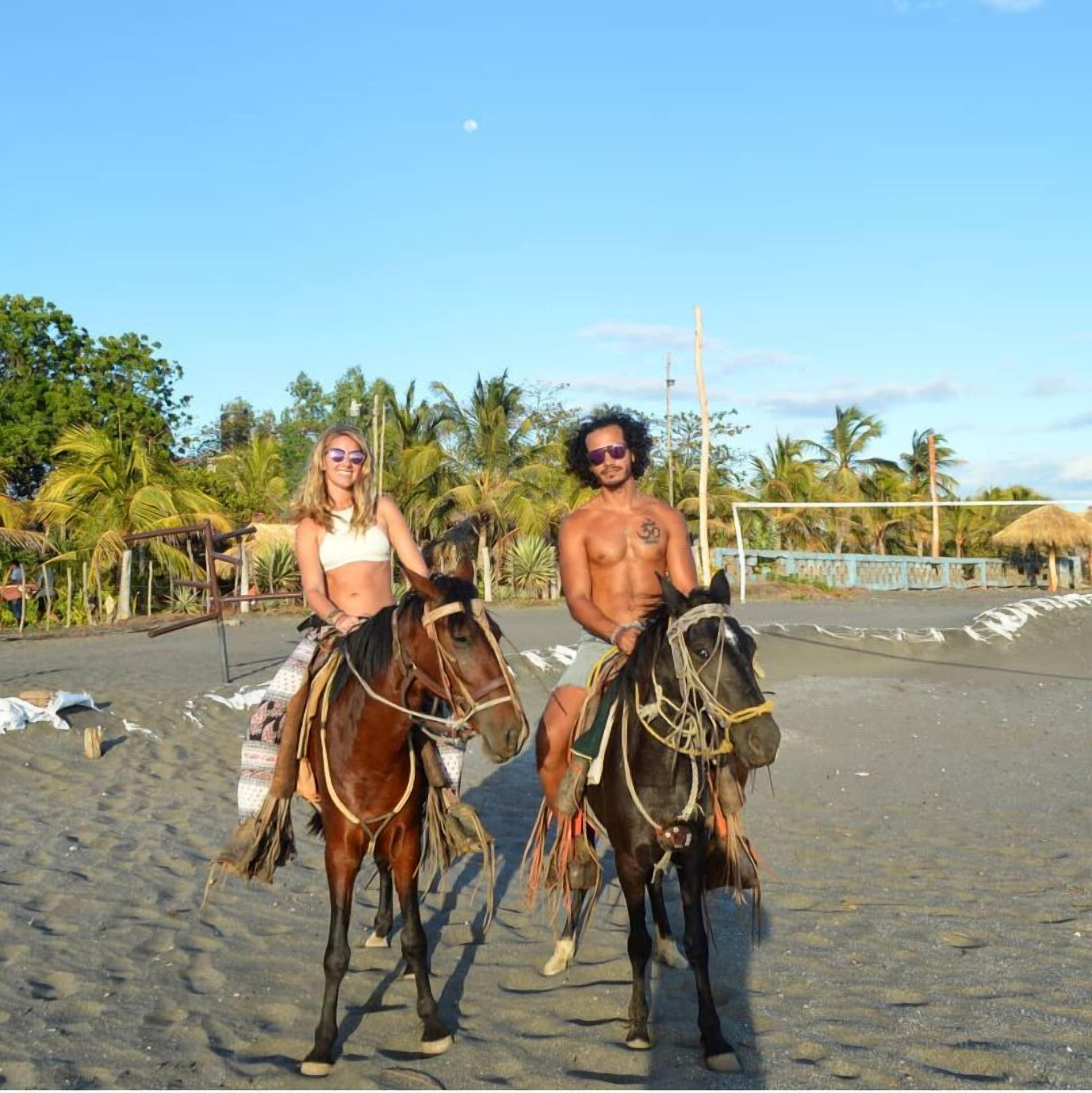 Deux personnes à cheval sur une plage, avec des palmiers et une structure à toit de chaume en arrière-plan.