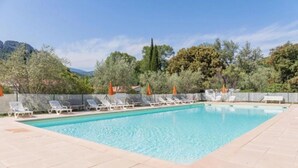 Una piscina rectangular con agua azul cristalina, rodeada de sillones blancos y sombrillas naranjas, con árboles y montañas como telón de fondo.