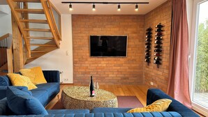 A modern living room with a blue sofa, a wooden staircase, a brick wall, a mounted TV, and a wine rack. - Wine and forest, cozy vacation home with outdoor sauna and a big garden (Landau in der Pfalz)