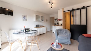 Een moderne woonkamer met een eethoek, een grijze bank, een aan de muur bevestigde televisie en een keuken met een magnetron en kastjes.