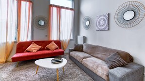 Una sala de estar moderna con un sofá rojo, un sofá gris y una mesa de centro blanca. Hay tres espejos en la pared y un radiador.