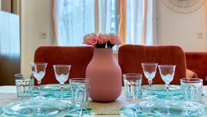 Una mesa de comedor dispuesta con cristalería transparente y un jarrón rosa con flores, y un sofá tapizado en rojo al fondo.