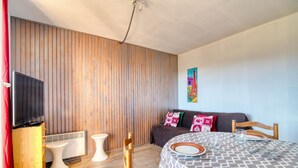 A modern room with a wooden slat wall, a flat-screen TV, a sofa with patterned cushions, a dining table set with plates and glasses, and a small white stool. - Le Cocon des 3 Domaines - Appt pour 4 (Ax-les-Thermes)