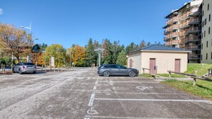 A parking area with two cars, a small building, and a multi-story residential building in the background. - Le Cocon des 3 Domaines - Appt pour 4 (Ax-les-Thermes)