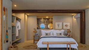 Een slaapkamer met een groot bed, een houten hoofdbord, nachtkastjes, een bankje en uitzicht op zee door een rond raam.