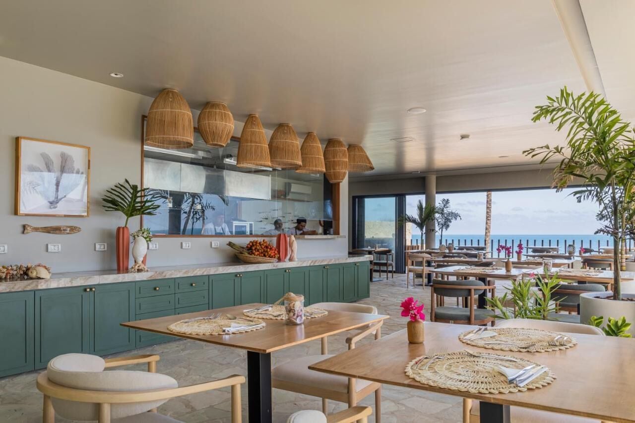 Un restaurant moderne offrant une vue sur la mer, avec des meubles verts, des tables en bois et des luminaires suspendus en fibres tressées.