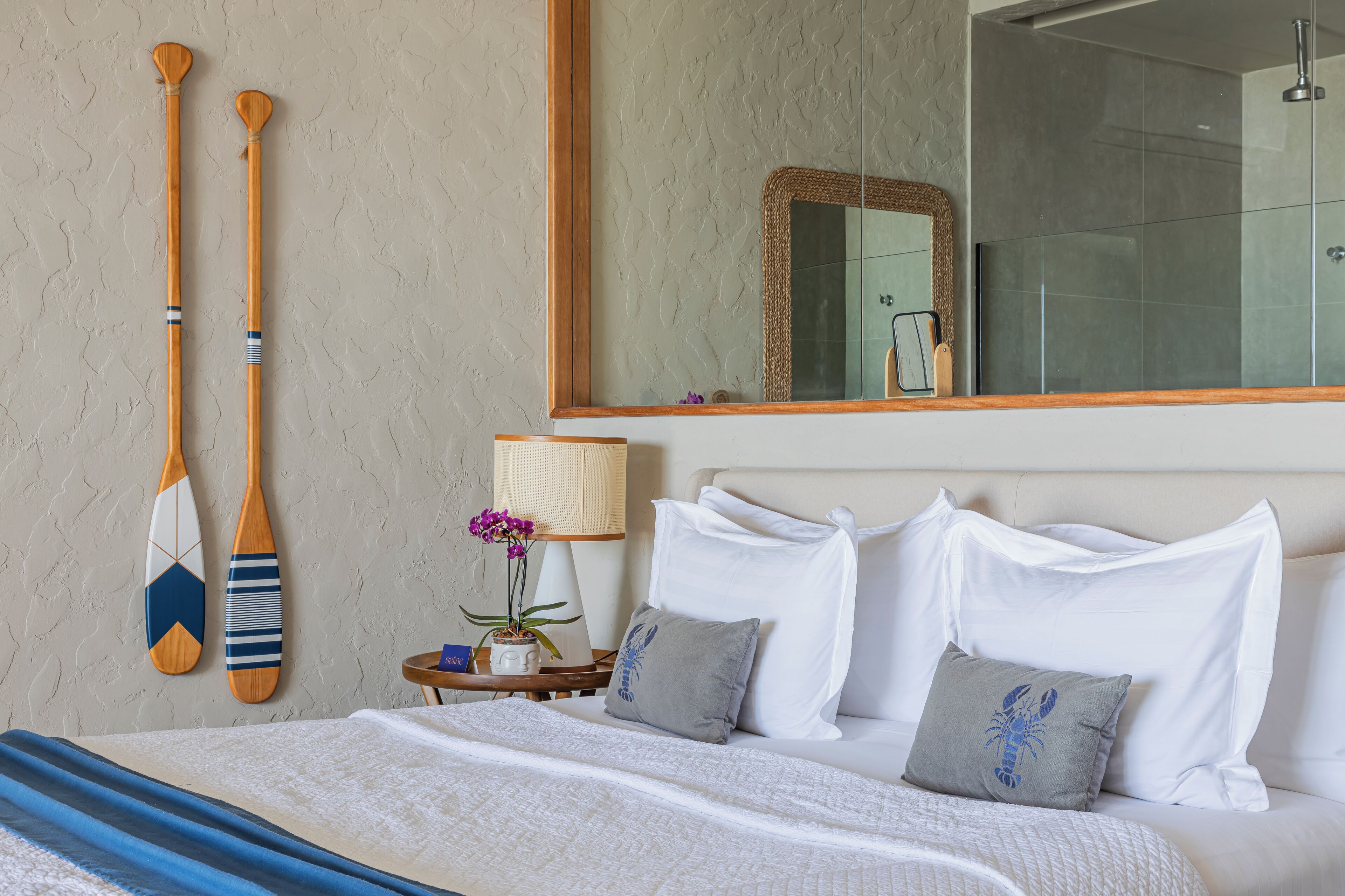 Quarto de hotel com cama, travesseiros, mesa de cabeceira com luminária, espelho e dois remos de madeira na parede.