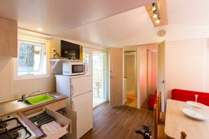 A compact kitchen with a microwave, sink, and stove, a small dining area with a red chair, and a view through an open door. - ZEcamping 3* 200m plage piscine animaux acceptés locatif TV BBQ clim (Calella)