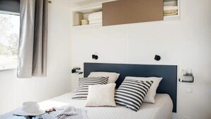 Une chambre à coucher bien rangée, avec un lit, des oreillers, une table de chevet, une fenêtre avec des rideaux et une étagère au-dessus du lit.