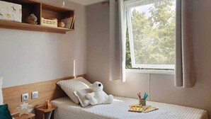 Une chambre à coucher bien rangée, avec un lit, une table de chevet, une étagère avec des livres et des objets décoratifs, et une fenêtre donnant sur des arbres.
