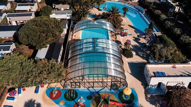 Eine Vogelperspektive zeigt einen Urlaubsort mit einem großen Schwimmbecken, einer überdachten Wasserrutsche und einem Spielbereich für Kinder.