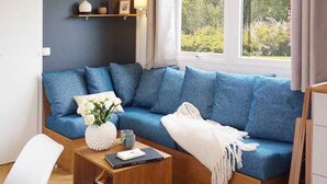 Ein modernes Wohnzimmer mit einem blauen Sofa, einem weißen Sessel und einem kleinen Holztisch. Über dem Sofa befinden sich Regale mit Dekoration, und ein Fenster bietet Blick auf Grünflächen.
