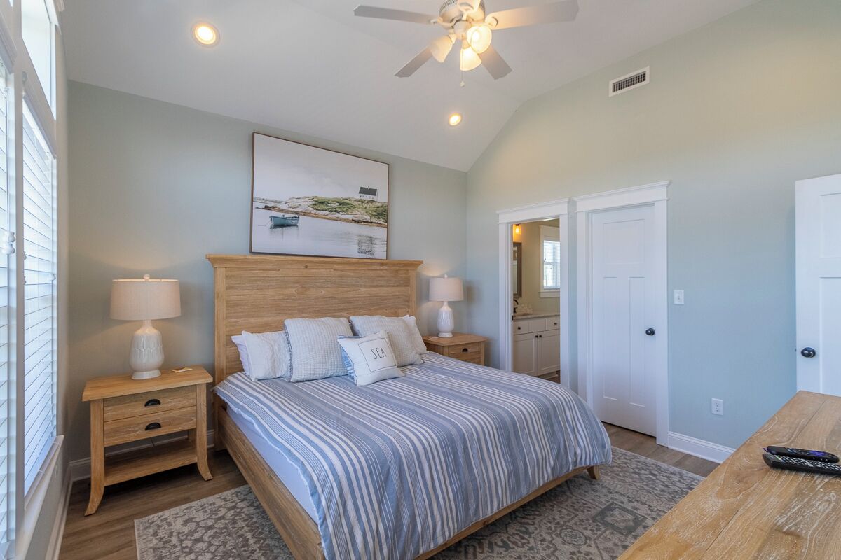 A bedroom with a large bed, two bedside tables, a ceiling fan, a nightstand with a lamp, a framed picture on the wall, and a wooden table with a remote control.