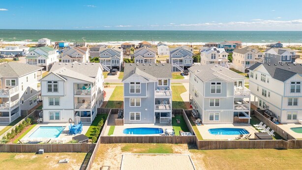 A coastal residential area with multiple two-story houses, each featuring a front yard with a swimming pool and a wooden fence. - Sun Realty gorgeous Kitty Hawk home with elevator, just steps from the beach (Kitty Hawk)
