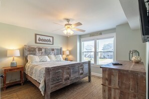 A bedroom with a wooden bed, two bedside tables, a ceiling fan, and a large window with blinds. - Sun Realty gorgeous Kitty Hawk home with elevator, just steps from the beach (Kitty Hawk)